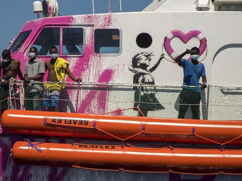 Ein Junge an Bord der «Louise Michel», das vom renommierten Künstler Banksy finanziert wird, winkt der Besatzung des Rettungsschiffes «Astral». Das Rettungsschiff «Louise Michel» kann sich nach Angaben der Besatzung mit mehr als 200 Migranten an Bord nicht länger sicher fortbewegen. Foto: Santi Palacios/AP/dpa