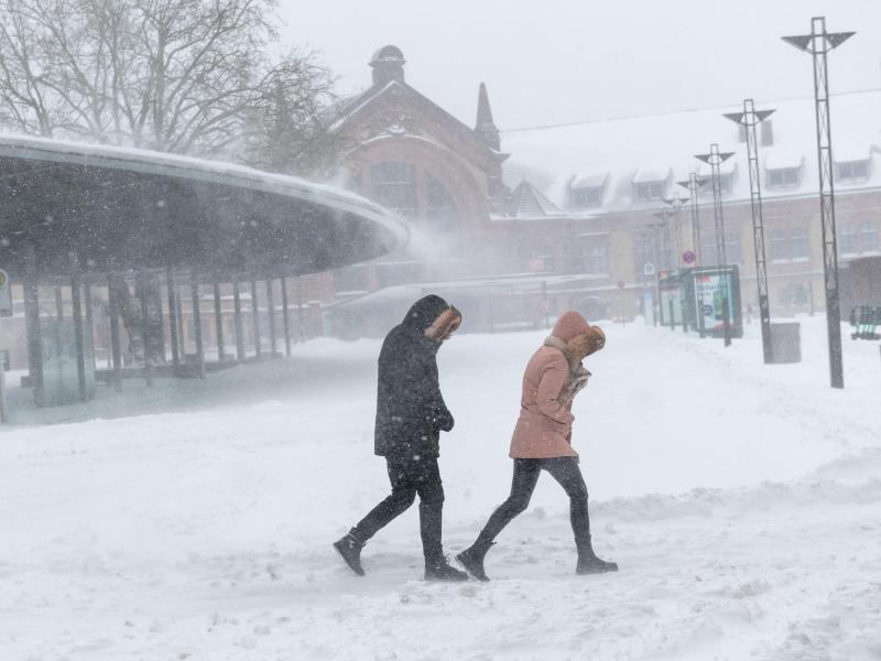 Zwei Menschen gehen mit eingezogenen Köpfen durch starken Schneefall mit Windböen vor dem Osnabrücker Hauptbahnhof. Foto: Jonas Walzberg/dpa