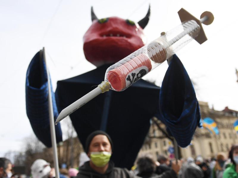 Ein Teilnehmer trägt auf der Demonstration in München eine Teufelspuppe mit Impfspritze. Foto: Angelika Warmuth/dpa