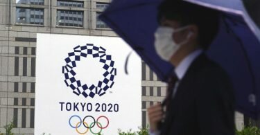 Ein Mann mit Mund-Nasen-Schutzt geht im Regen an einem Banner der Olympischen Spiele Tokio 2020 vorbei. Foto: Eugene Hoshiko/AP/dpa