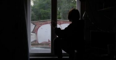 Eine Jugendliche sitzt in einem abgedunkelten Zimmer am Fenster. Foto: Paul Zinken/dpa