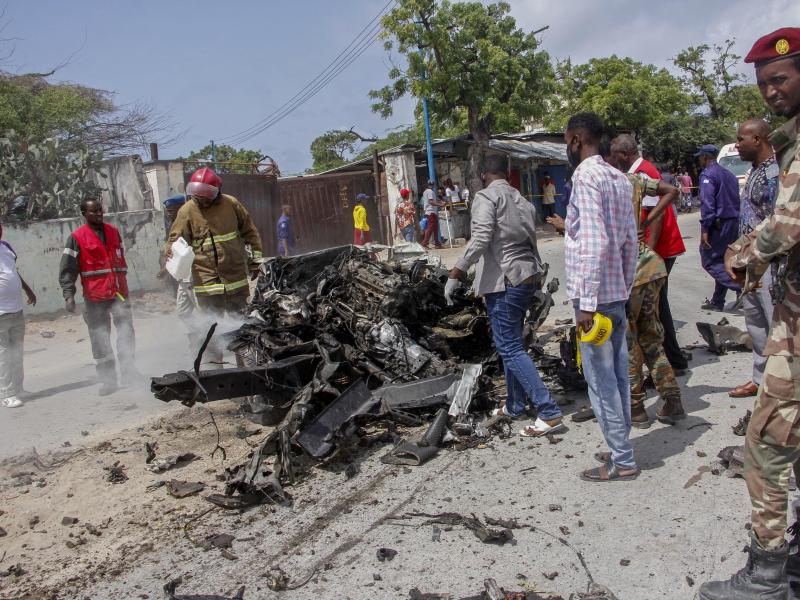 Sicherheitskräfte und Zivilisten stehen in der Nähe eines Autowracks nach dem ersten Anschlag im somalischen Mogadischu. Die islamistische Terrormiliz Al-Shabaab hat nun sechs weitere Mörsergeschosse abgefeuert. Foto: Farah Abdi Warsameh/AP/dpa
