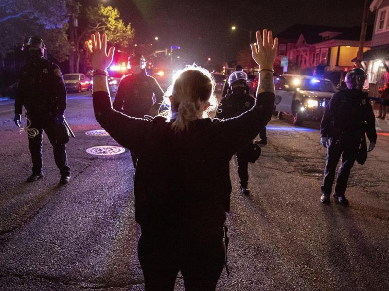 Eine Demonstrantin steht bei einem Protest gegen Polizeigewalt in Oakland mit hochgestreckten Armen vor Polizisten. Foto: Ethan Swope/AP/dpa