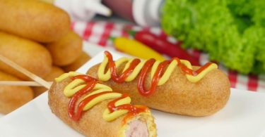 Klassische Corn Dogs bestehen aus aufgespießten Hot-Dog-Würstchen in einer Maisteighülle. Traditionell gibt es Senf und Ketchup dazu. Foto: amerkanisch-kochen.de/dpa-tmn