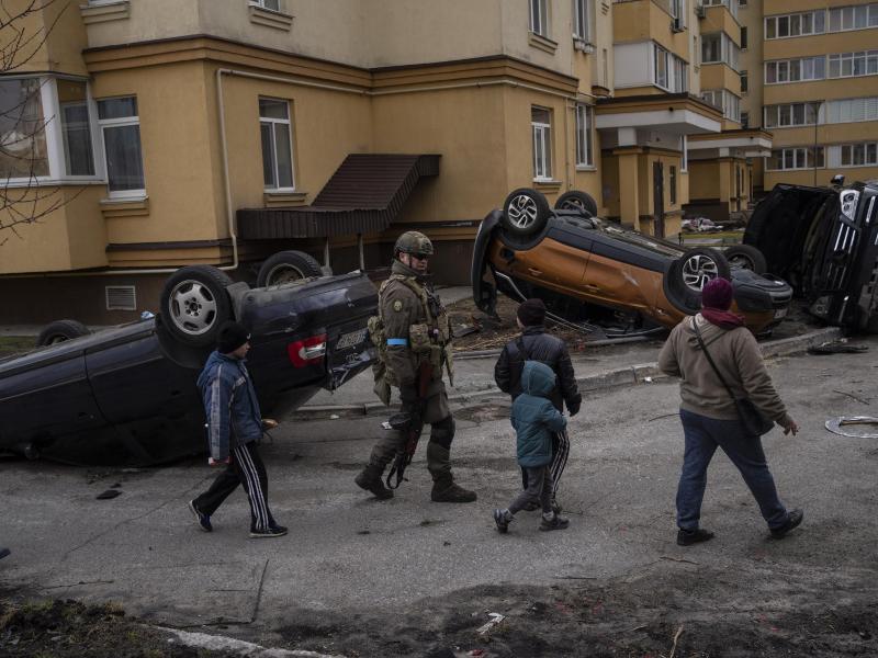 Ein ukrainischer Soldat geht in Butscha mit Kindern an zerstörten Autos vorbei. Foto: Rodrigo Abd/AP/dpa