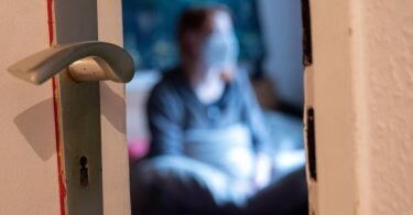 Eine Frau sitzt während der Isolation nach einem positiven Corona-PCR-Test auf ihrem Bett. Foto: Sebastian Gollnow/dpa