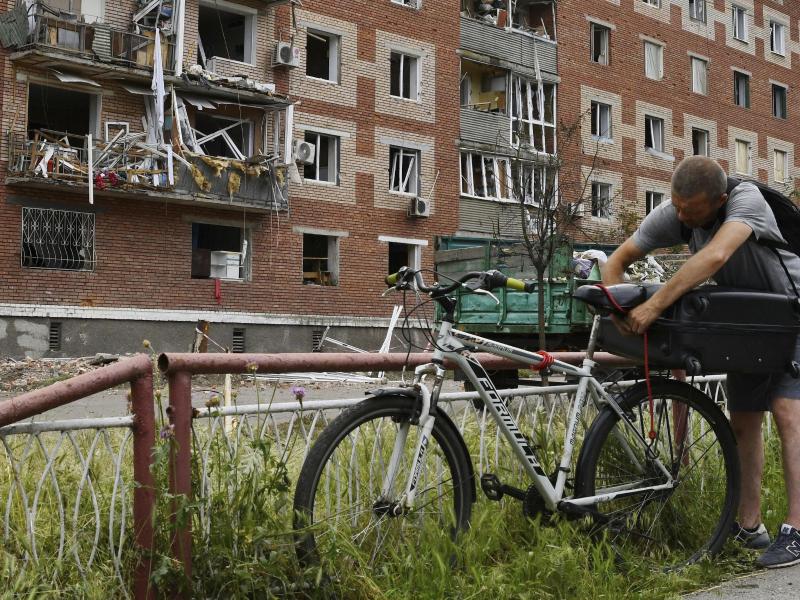 Ein Mann in Slowjansk zurrt einen Koffer auf seinem Fahrrad fest. Das Wohnhaus hinter ihm wurde bei einem nächtlichen Raketeneinschlag beschädigt. Foto: Andriy Andriyenko/AP/dpa